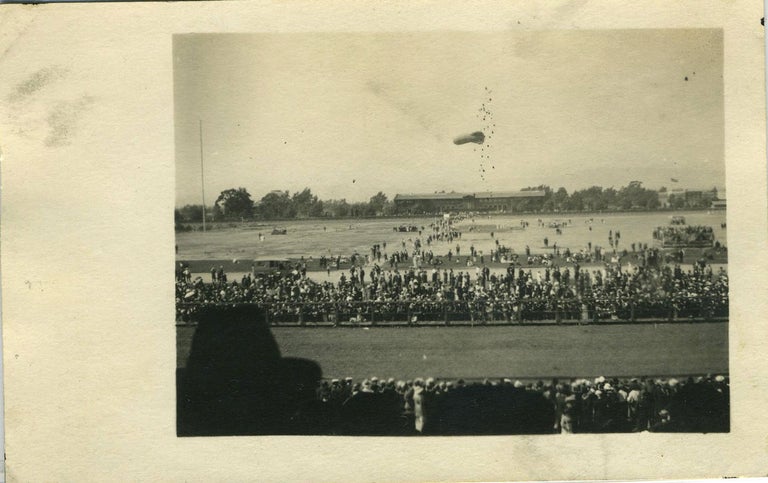 Item #19402 Military event/gathering, perhaps at a race track, with a small airship flying, it's tail end surrounded both above and below with a circle of projectiles. Real photo postcard.